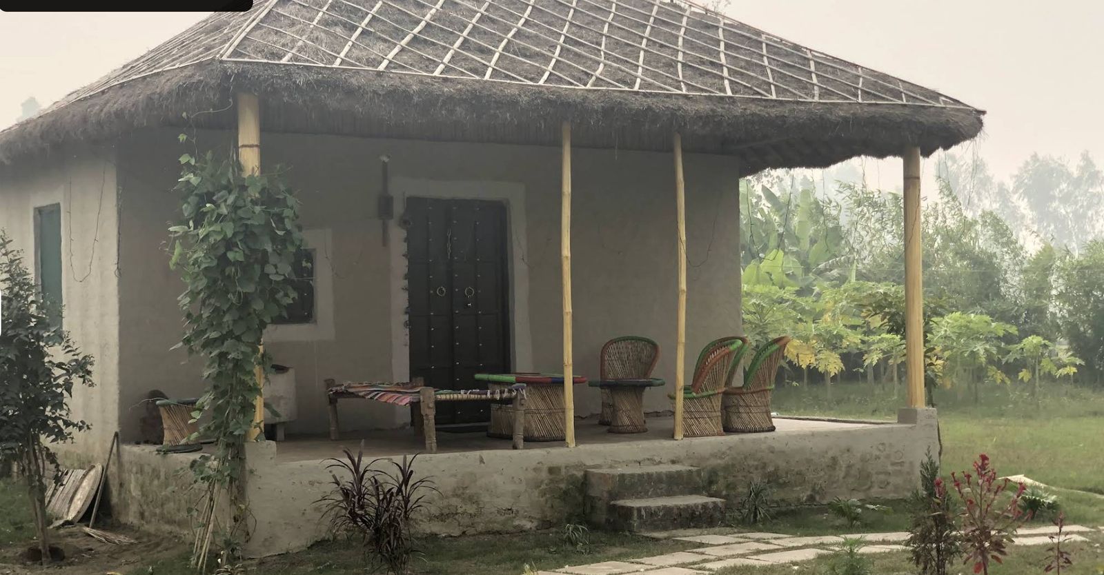 Mud-plastered thatched cottage with a wraparound porch on a misty morning