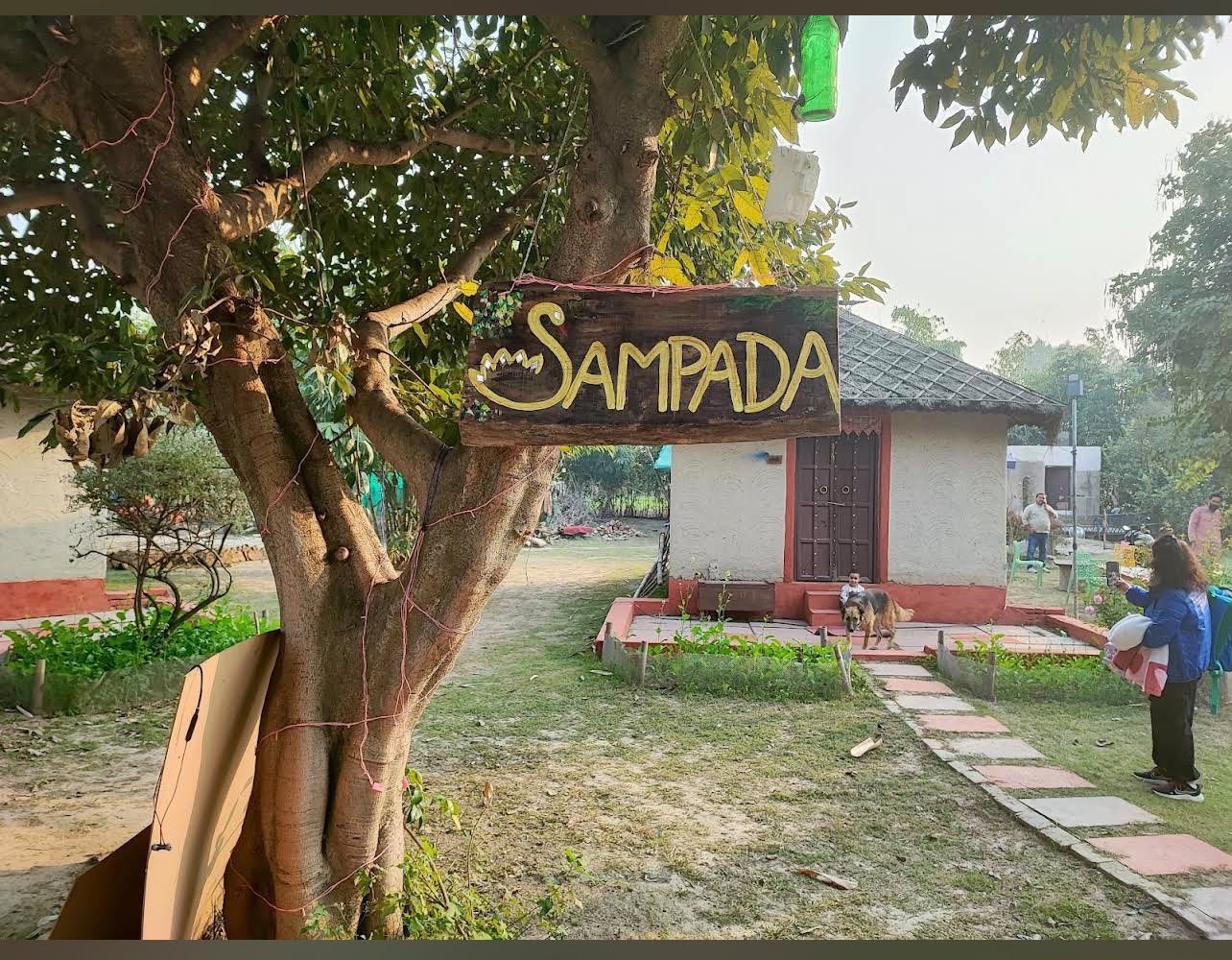 Hand-painted Sampada signboard hanging from a tree at the entrance, with a cottage and dog in the background